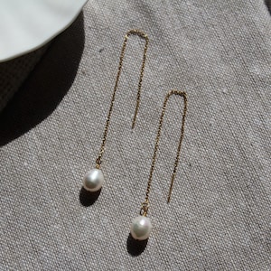 May include: A pair of gold-coloured threader earrings. Each earring has a small, lustrous white pearl hanging from a fine chain. The earrings are displayed on a textured, grey surface.