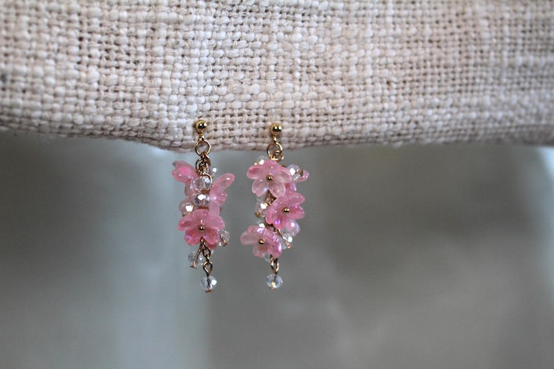 May include: A pair of delicate earrings featuring pink flower-shaped charms dangling from gold-toned posts. The charms are arranged in a cascading design, creating a whimsical and feminine look.