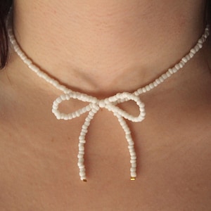 May include: A white beaded choker necklace with a bow in the centre. The bow is made of the same white beads and has two long strands that hang down the front of the neck.