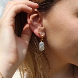 May include: A close-up of a gold hoop earring with a small, round stud earring. The hoop earring features a light grey, rectangular-shaped pearl pendant. A person's hand is touching their ear.