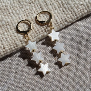 May include: Gold-coloured hoop earrings featuring three dangling white star charms. The earrings are set against a textured, beige fabric background. Each star charm is connected by a small gold bead.