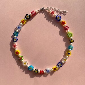 May include: A colorful flower necklace with white pearl beads. The necklace is made of glass and features a variety of colors, including red, yellow, blue, green, and purple.
