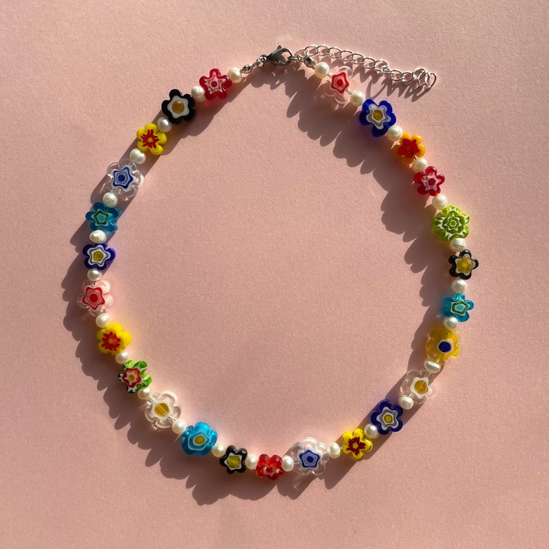 May include: A colorful flower necklace with white pearl beads. The necklace is made of glass and features a variety of colors, including red, yellow, blue, green, and purple.