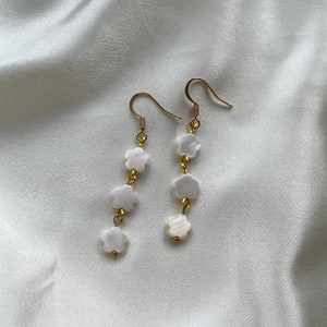 Può includere: Un paio di orecchini dorati con perline bianche a forma di fiore. Ogni orecchino ha quattro perline.