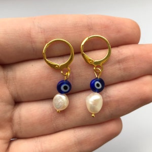 May include: Gold-colored leverback earrings. Each earring features a blue bead with an eye design, a white pearl, and a small gold bead. The earrings are displayed against a light background.