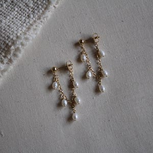 May include: A pair of gold earrings with hanging white pearls. The earrings have a post-back design and feature multiple strands of delicate gold chains, each adorned with small, round pearls. The earrings are displayed on a neutral-toned fabric.