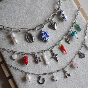 May include: Three silver chain necklaces with assorted charms. Charms include pearls, a seashell, a heart, a chilli pepper, a star, a horseshoe, and a cherry. The necklaces are displayed on a light beige surface.