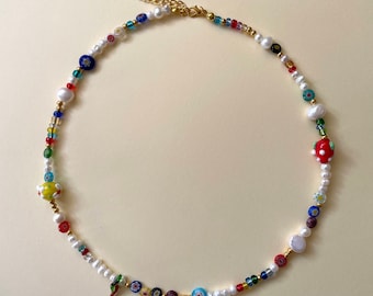 May include: A colorful beaded necklace featuring a variety of beads, including white pearls, small colorful glass beads, and decorative glass beads with floral and other designs. The necklace has a gold-colored chain and clasp.