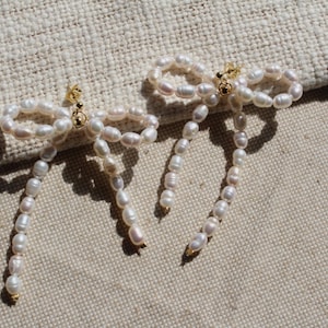 May include: A pair of pearl earrings with a bow design. The earrings are made of white pearls and have a gold-tone finish.