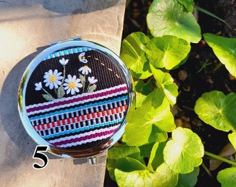 May include: A round, silver-framed compact mirror with a black fabric base. The fabric is decorated with embroidered white flowers and a bee, and has a colorful striped pattern.
