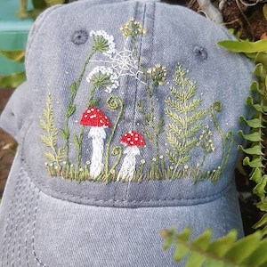 Può includere: Un berretto da baseball grigio con un ricamo a forma di funghi, felci e fiori. Il ricamo è in tonalità di verde, bianco e rosso.