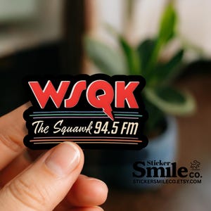 May include: A black sticker with the text "WSQK The Squawk 94.5 FM" in red and white. The sticker has a retro design with colorful horizontal lines. The sticker is held by a person.