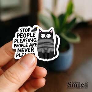 May include: A die-cut sticker featuring a black cat with wide eyes wearing a gray and black striped sweater. Next to the cat is a small black bird. The text reads: STOP PEOPLE PLEASING. PEOPLE ARE NEVER PLEASED. The sticker has a white border.