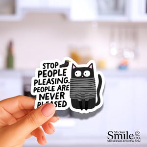 May include: A die-cut sticker featuring a black cat with wide eyes wearing a gray and black striped sweater. A small black bird sits above the cat. The text reads: STOP PEOPLE PLEASING. PEOPLE ARE NEVER PLEASED. The sticker has a white border.