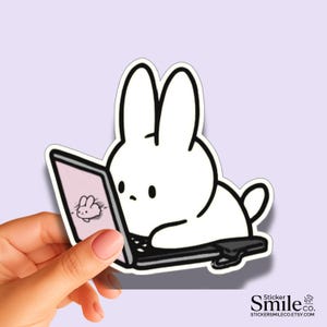 May include: A white rabbit sticker with long ears and a small tail, depicted sitting and looking at a black laptop screen. The screen displays a small graphic of a rabbit. The sticker features a thick white border and bold black outlines.