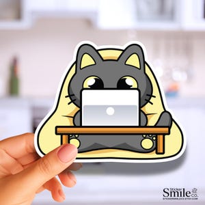 May include: A die-cut sticker featuring a cartoon dark gray cat with large eyes sitting at a brown wooden desk while using a silver laptop. The cat is positioned against a pale yellow background, and the text Sticker Smile co. is visible.
