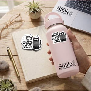 May include: A light pink insulated water bottle featuring a white sticker with a black cat wearing a grey striped sweater and the text: STOP PEOPLE PLEASING. PEOPLE ARE NEVER PLEASED. A matching sticker is placed on a notebook next to the bottle.