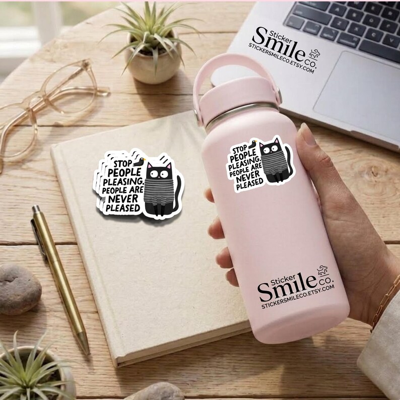 May include: A light pink insulated water bottle featuring a white sticker with a black cat wearing a grey striped sweater and the text: STOP PEOPLE PLEASING. PEOPLE ARE NEVER PLEASED. A matching sticker is placed on a notebook next to the bottle.