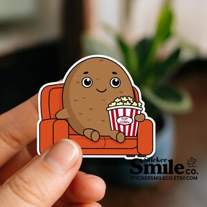 May include: A sticker featuring a cartoon potato character with a smiling face, seated on an orange couch, holding a popcorn bucket. The popcorn bucket has red and white stripes and the word "Popcorn". The sticker has a white border.