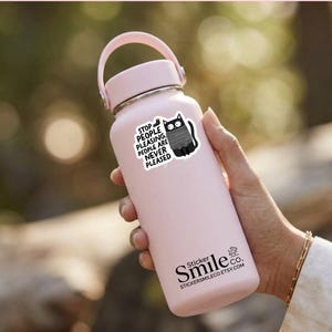 May include: A light pink insulated water bottle with a handle on the lid. A sticker on the bottle features a black cat wearing a grey sweater and the text: STOP PEOPLE PLEASING. PEOPLE ARE NEVER PLEASED. The bottle also displays the text Sticker Smile co.