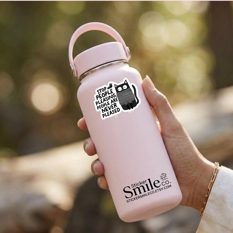 May include: A light pink insulated water bottle with a handle on the lid. A sticker on the bottle features a black cat wearing a grey sweater and the text: STOP PEOPLE PLEASING. PEOPLE ARE NEVER PLEASED. The bottle also displays the text Sticker Smile co.