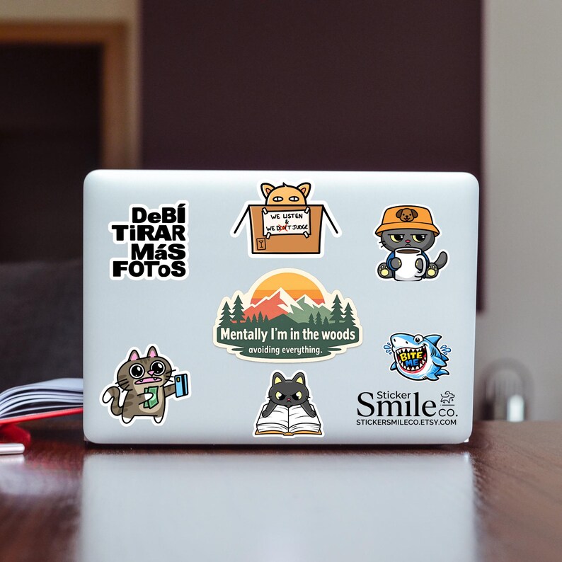 May include: A white laptop lid covered in various stickers including a cat in a box, a cat in a bucket hat with a mug, a shark with the text BITE ME, a cat reading a book, a mountain landscape with the text Mentally I'm in the woods, and text.