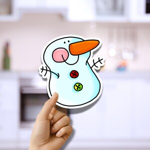May include: A cartoon snowman sticker with a light blue body, orange carrot nose, and red and green button details. The snowman has a pink blush and black eyes. The sticker is held up by a hand.