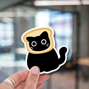 May include: A die-cut sticker featuring a solid black cat with large white circular eyes and whiskers, framed by a light tan slice of bread surrounding its head. The sticker has a thick white border and a smooth, matte-like finish.