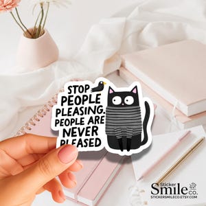 May include: A die-cut sticker featuring a black cat wearing a gray and black striped sweater. The cat has wide eyes and is next to a small black bird. The text reads: STOP PEOPLE PLEASING. PEOPLE ARE NEVER PLEASED. The sticker has a white border.