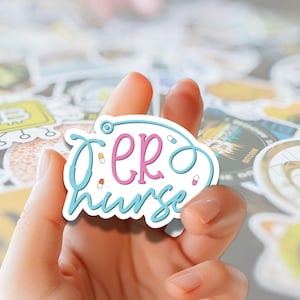 May include: A sticker with the words "ER nurse" in pink and blue, with a stethoscope design. The sticker also features small pill illustrations. The sticker is held in a hand, with other stickers visible in the background.