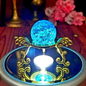 K&ouml;nnte beinhalten: Eine blaue Glaskugel mit glitzerndem, rissigem Eiseffekt sitzt auf einem goldenen Metallst&auml;nder mit verzierten Details. Der St&auml;nder steht auf einer drehbaren Plattform mit einer verspiegelten Oberfl&auml;che.