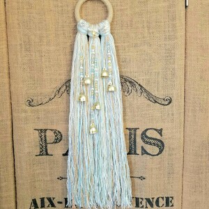 May include: A white and light blue yarn tassel wall hanging with gold bells. The tassel is hanging from a wooden ring.
