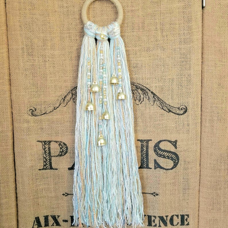 May include: A white and light blue yarn tassel wall hanging with gold bells. The tassel is hanging from a wooden ring.