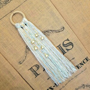 May include: A light blue and white macrame wall hanging with gold bells. The hanging is made with a wooden ring and has a fringe at the bottom.
