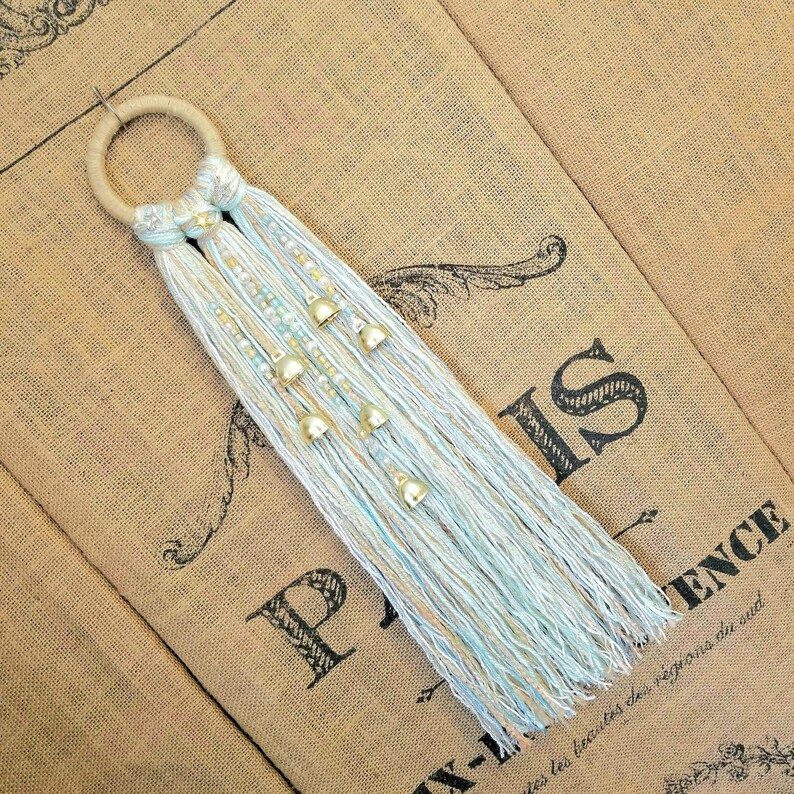 May include: A light blue and white macrame wall hanging with gold bells. The hanging is made with a wooden ring and has a fringe at the bottom.