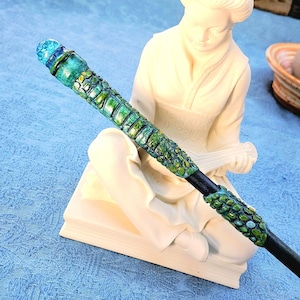 May include: A long, slender wand featuring a textured, green and black scale-like pattern along the handle and shaft. The top is capped with a translucent, spherical blue orb. The wand has a smooth, dark, tapered end extending from the textured grip.