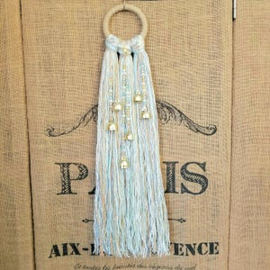 May include: A macrame wall hanging with a hoop and tassels in shades of blue and white. The tassels are adorned with small gold bells.