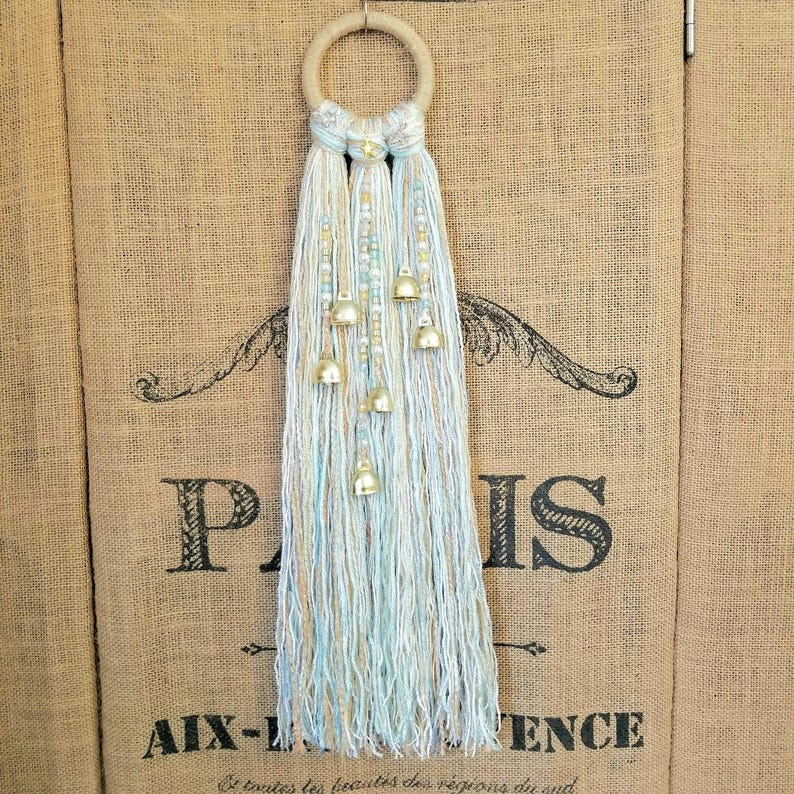 May include: A macrame wall hanging with a hoop and tassels in shades of blue and white. The tassels are adorned with small gold bells.