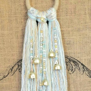 May include: A light blue and white yarn tassel wall hanging with gold bells and a star charm. The tassel is made of multiple strands of yarn and beads.