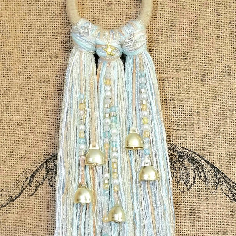 May include: A light blue and white yarn tassel wall hanging with gold bells and a star charm. The tassel is made of multiple strands of yarn and beads.