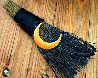 May include: A black broom with a black twine handle and a gold crescent moon charm. The broom is lying on a wooden surface.
