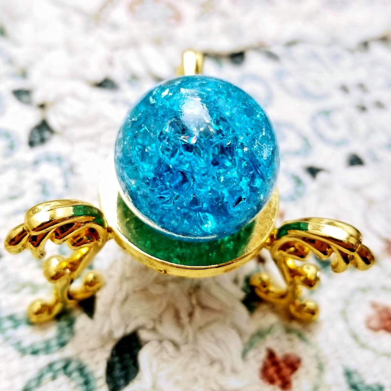 K&ouml;nnte beinhalten: Eine blaue Glaskugel mit glitzernder, gesprenkelter Textur sitzt auf einem goldenen St&auml;nder mit drei Beinen. Der St&auml;nder hat ein dekoratives, verziertes Design.