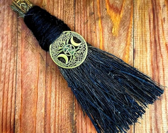 May include: A small broom made of dark, thin twigs bound with thick black yarn. A circular gold-toned metal charm featuring a central pentagram surrounded by three crescent moons and intricate Celtic knotwork is attached to the handle area.