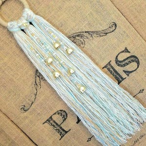 May include: A light blue and beige macrame wall hanging with gold bells. The hanging is made with a variety of textures and is a beautiful addition to any home decor.