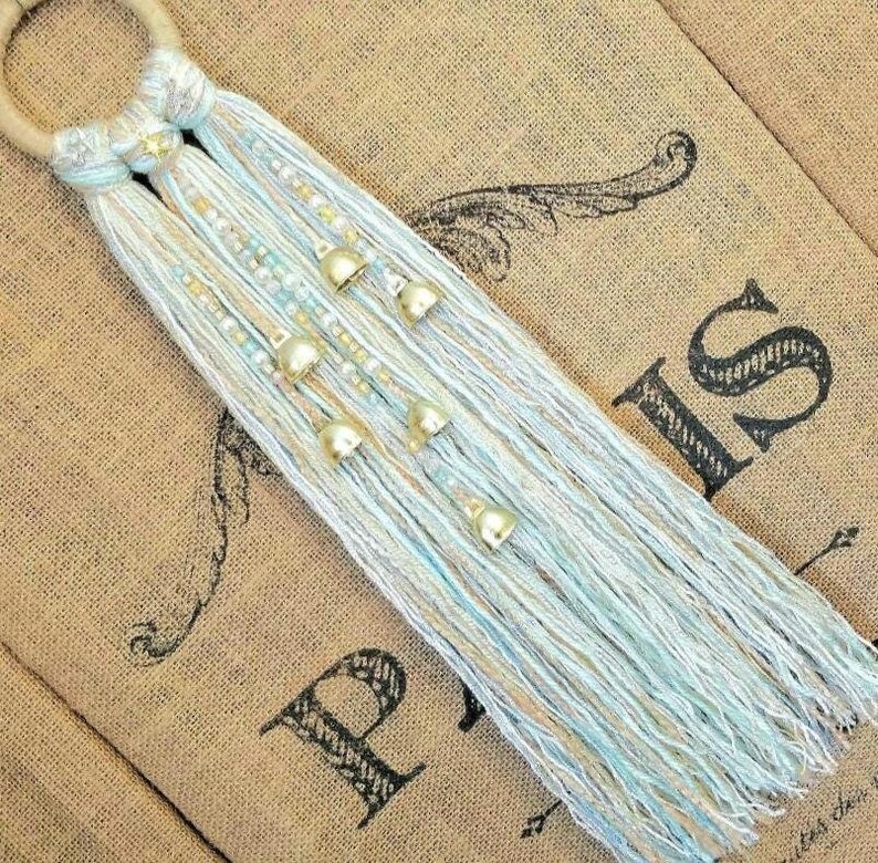 May include: A light blue and beige macrame wall hanging with gold bells. The hanging is made with a variety of textures and is a beautiful addition to any home decor.