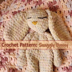 以下が含まれることがあります： ピンクと黄色の色合いの毛糸で編まれたぬいぐるみウサギが、ピンクと白の毛糸で編まれた毛布の上に横たわっています。おもちゃは、目が閉じられていて、小さな黄色い鼻があるシンプルな顔をしています。「Crochet Pattern: Snuggly Bunny」というテキストがおもちゃの上に表示されています。画像には、Rowan Tree FiberArtのロゴが含まれています。