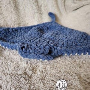 Puede incluir: Un gorro de tiburón de ganchillo azul hecho a mano con una aleta y ribete blanco. El gorro está hecho de hilo suave y texturizado y está diseñado para la cabeza de un niño. El gorro se muestra sobre una superficie gris y difusa.