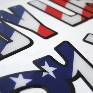 Puede incluir: Primer plano de una pegatina 3D con la palabra "USA" en letras con contorno negro. Las letras est&aacute;n rellenas con un patr&oacute;n de bandera estadounidense rojo, blanco y azul.