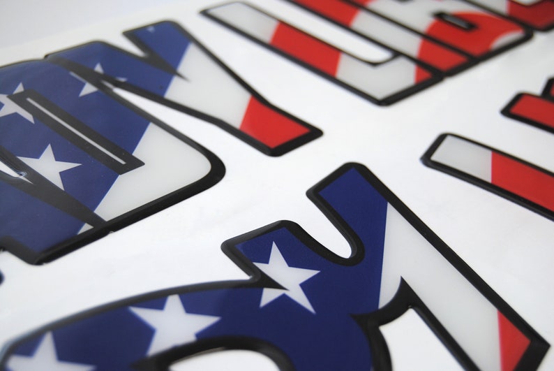 Puede incluir: Primer plano de una pegatina 3D con la palabra "USA" en letras con contorno negro. Las letras est&aacute;n rellenas con un patr&oacute;n de bandera estadounidense rojo, blanco y azul.