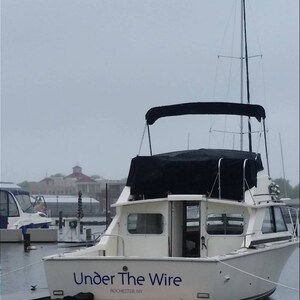 K&ouml;nnte beinhalten: Ein wei&szlig;es Boot, das an einem Steg in einer Marina liegt, mit einem schwarzen Stoffverdeck und dem Schriftzug "Under The Wire Rochester, NY" an der Seite.